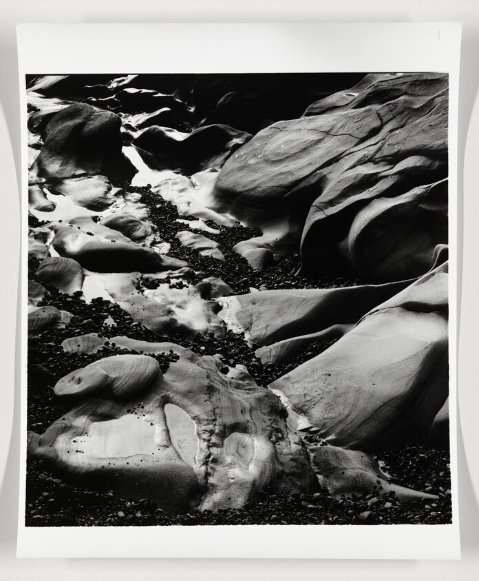 A black and white photograph depicting a rocky stream bed with smooth, eroded stones and pebbles. The contrasting light and shadows highlight the textures and patterns on the rock surfaces.