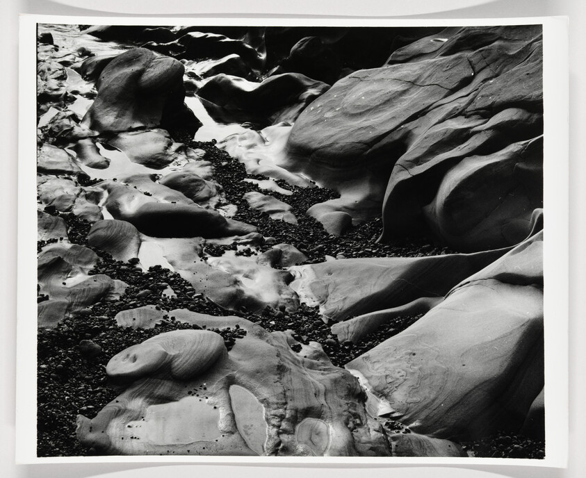 A black and white photograph of a rocky beach with smooth, eroded stones and pebbles scattered throughout. The textures and patterns on the rocks are highlighted by the play of light and shadow.