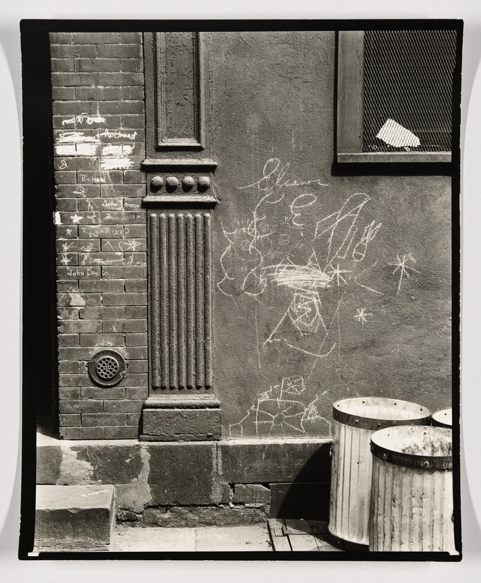 A brick and plaster alley wall with children's chalk drawings beside two metal trash cans.