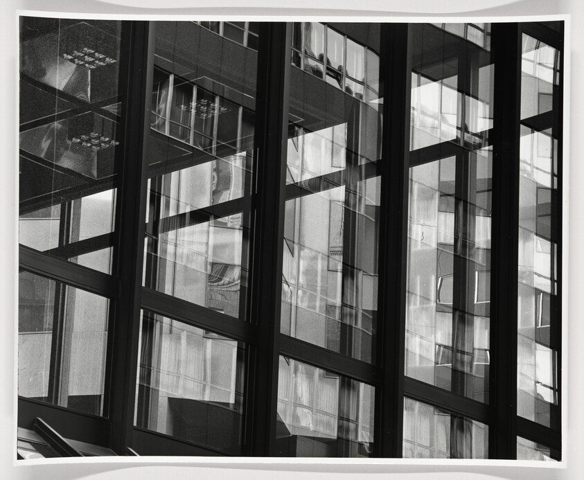 Tall glass windows with dark metal frames reflecting neighboring building facades in overlapping patterns.
