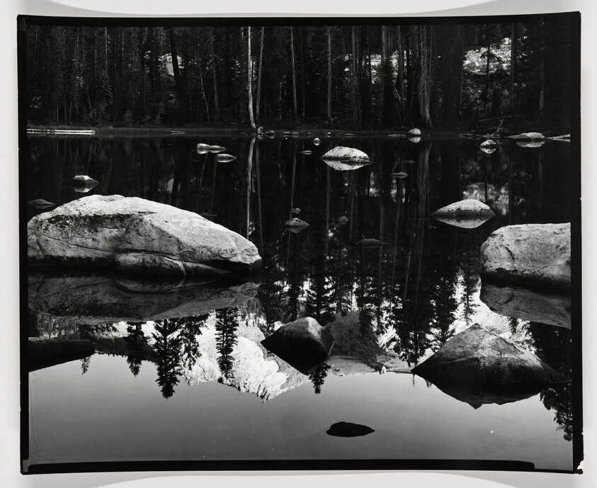A black and white photograph capturing a serene lake with a mirror-like reflection. Large rocks are scattered across the foreground, partially submerged, with their reflections creating a symmetrical effect. A dense forest lines the far shore, its trees also reflected in the still water, creating a tranquil and almost surreal landscape.