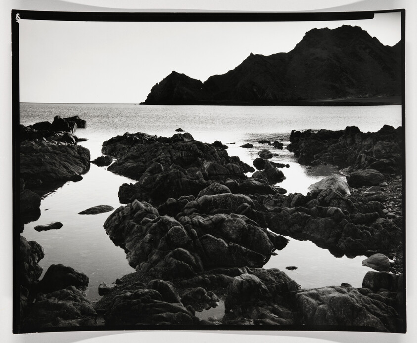 Black and white photograph of a rugged coastline with rocky formations in the foreground leading to a calm sea, with a silhouette of a mountain range in the background under a clear sky. The photo has a white border, and the top left corner appears to be slightly folded over.