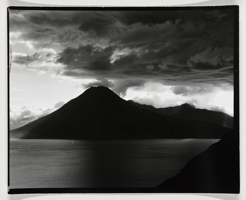 A black and white photograph capturing a dramatic landscape with a prominent mountain peak in the center, surrounded by a textured sky filled with clouds and a calm body of water in the foreground. Visible light rays break through the clouds, highlighting the mountain's silhouette and the serene water surface. The image has a vintage feel with a black border framing the scene.