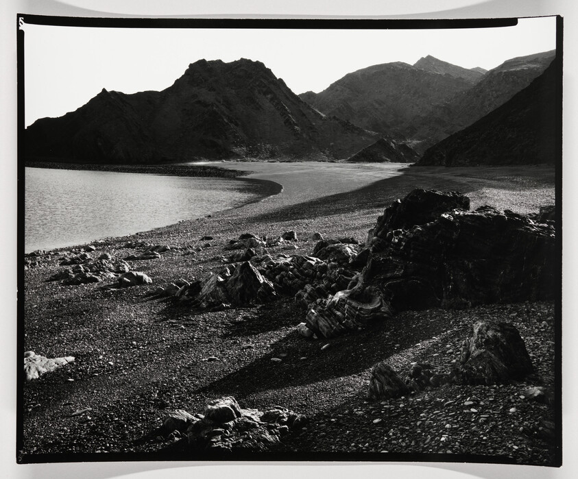 A black and white photograph depicting a rugged landscape with a curving shoreline on the left, leading to a range of layered mountains in the background. The foreground is strewn with rocks and pebbles, casting shadows in the low light, suggesting either early morning or late afternoon.