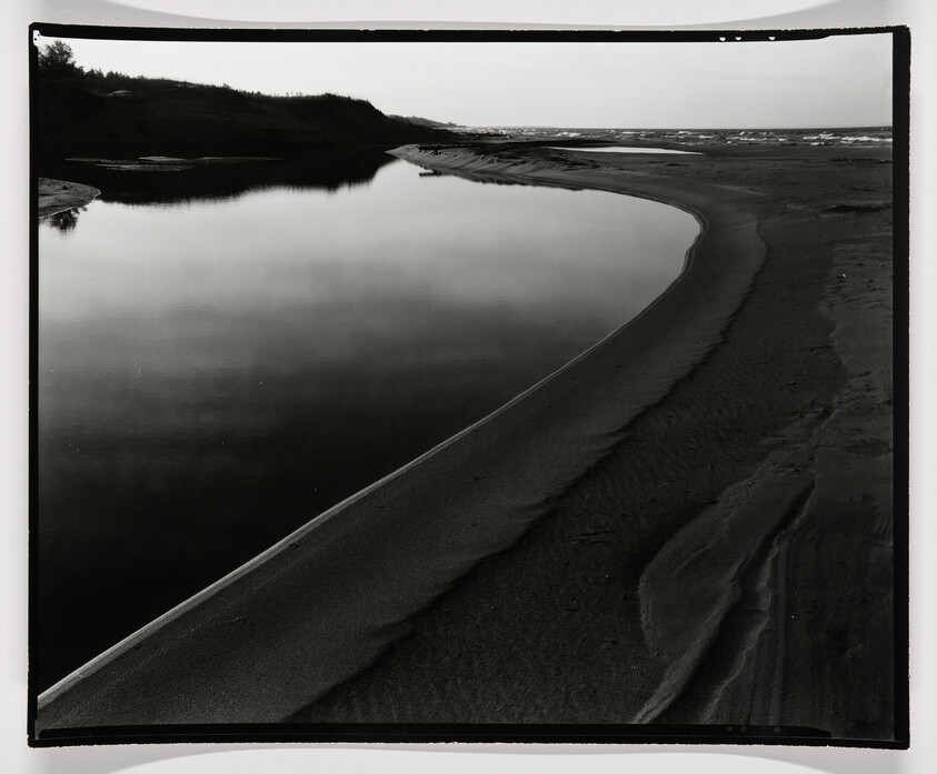 A black and white photograph capturing a serene coastal scene with a calm body of water reflecting the sky, bordered by a curving sandy shore with gentle ripples in the sand, leading to dunes and vegetation in the background. The horizon is visible in the distance where the water meets the sky.