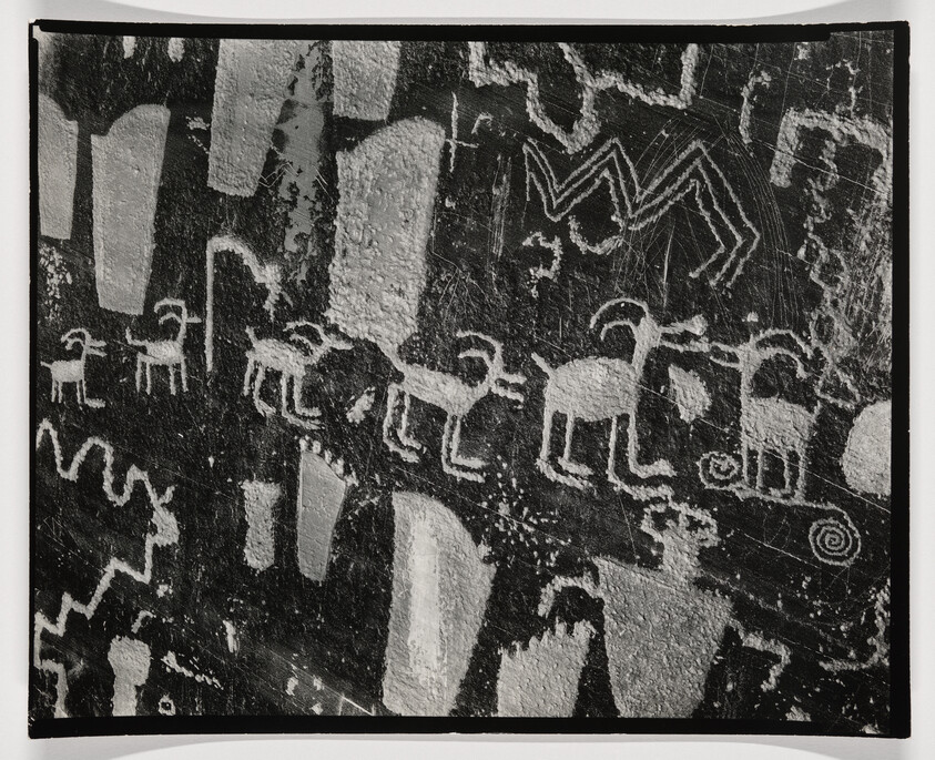 A black and white photograph of ancient rock art featuring a series of petroglyphs. The image shows various etched figures, including a procession of animals that resemble deer or elk, and abstract symbols such as spirals and zigzag patterns. The petroglyphs are set against a textured rock surface with visible weathering and scratches.