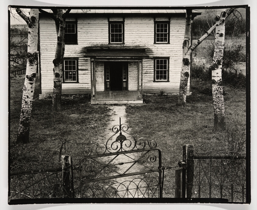 Old two-story wooden house with open iron gate and path leading to the front porch.