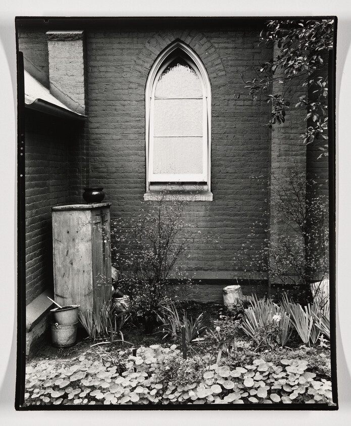 Arched window above a small garden bed with a weathered cabinet and metal pots.