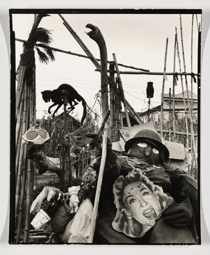 Scarecrow-like figures and eerie animal sculptures arranged in a bamboo frame, one holding a screaming woman's mask.