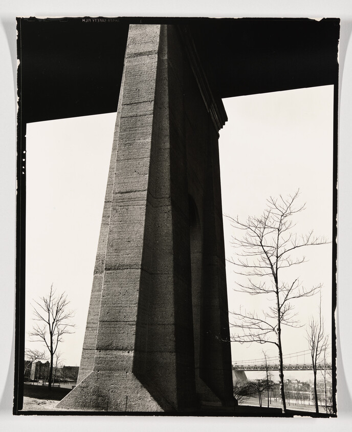 Massive concrete bridge pillar rising beside leafless trees with a distant suspension bridge.