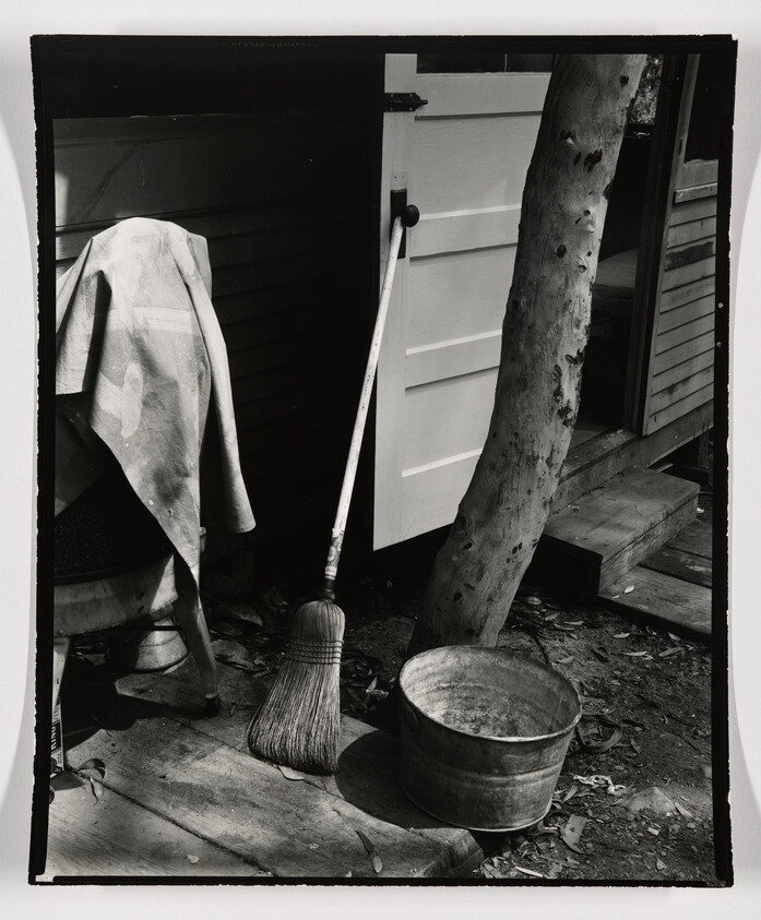 A broom leans against a doorway beside a metal bucket and a tree trunk.