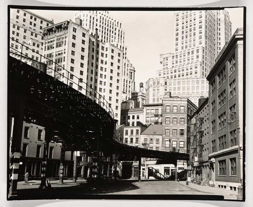 Curving elevated train tracks pass above a quiet city street lined with tall office buildings.