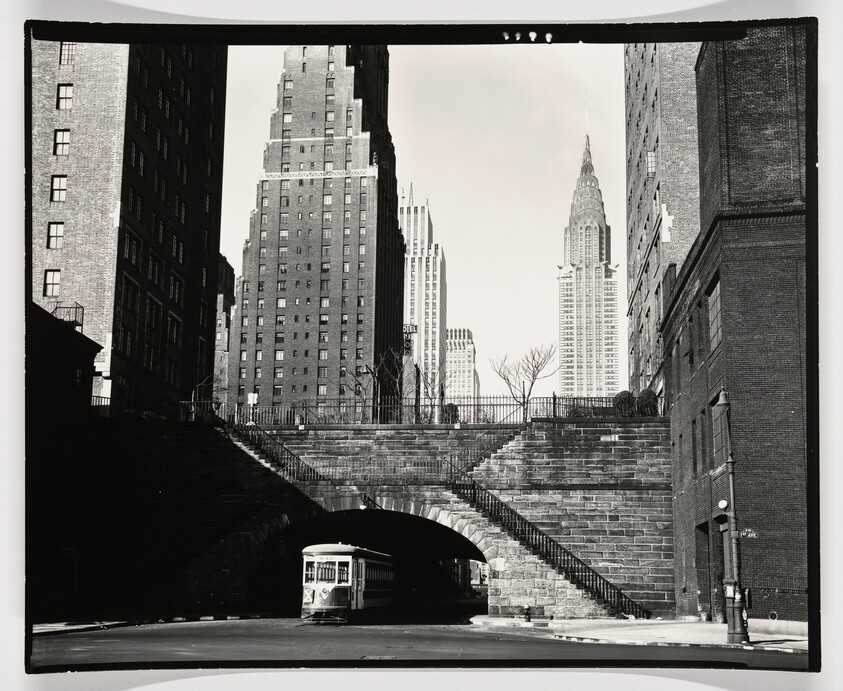 A streetcar emerges from a stone arch underpass with tall city skyscrapers rising behind it.