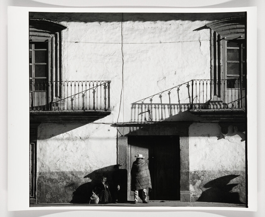 A black and white photograph capturing the facade of an old building with two balconies casting shadows on the wall. A person wearing a brimmed hat and a patterned shawl stands in the doorway, partially in sunlight and shadow, while another figure sits on the ground to the left, obscured in shadow.