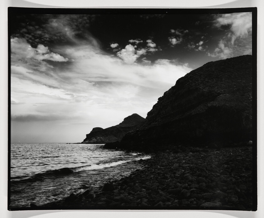 A black and white photograph depicting a dramatic coastal scene with a rocky shoreline in the foreground, gentle waves lapping at the beach, and rugged hills rising in the background under a sky filled with expressive clouds.