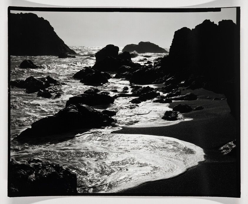A black and white photograph of a rugged coastline with waves crashing against rocks and washing over a sandy beach. The sunlight reflects off the water, creating a high-contrast scene with the dark rocks and shadows.