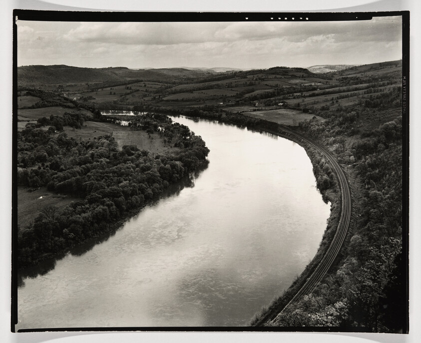 A wide river winds through rolling hills with railroad tracks following the riverbank.