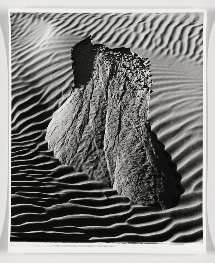 A black and white photograph capturing the stark contrast between the smooth, rippled texture of sand dunes and the rough, jagged surface of a large rock formation protruding from the sand. The play of light and shadow accentuates the patterns and textures in the scene.