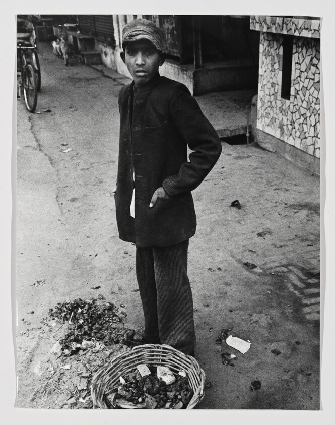 Young boy stands on a street next to a basket of collected debris, looking toward the camera.