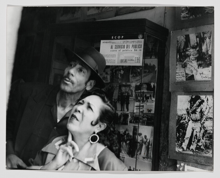 Man in a hat and woman with earrings look upward together near a wall of photographs.