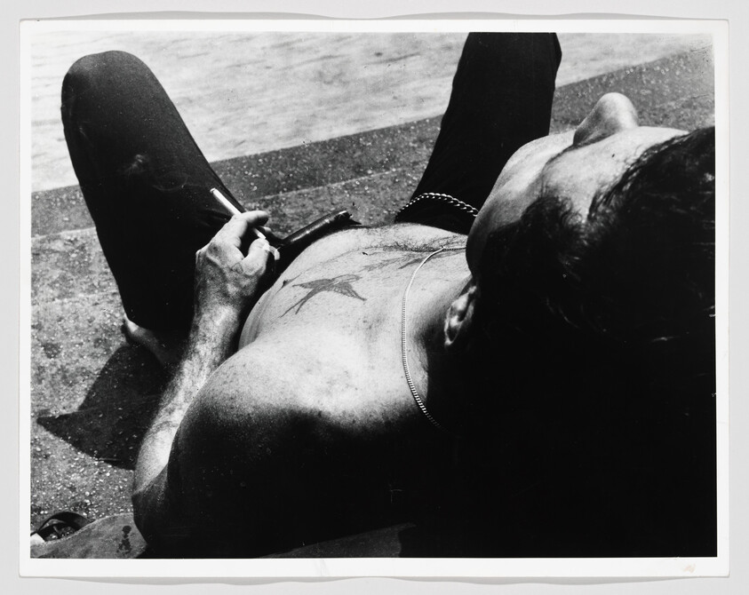 A shirtless man lies back smoking a cigarette, showing a star tattoo on his chest and a chain necklace.