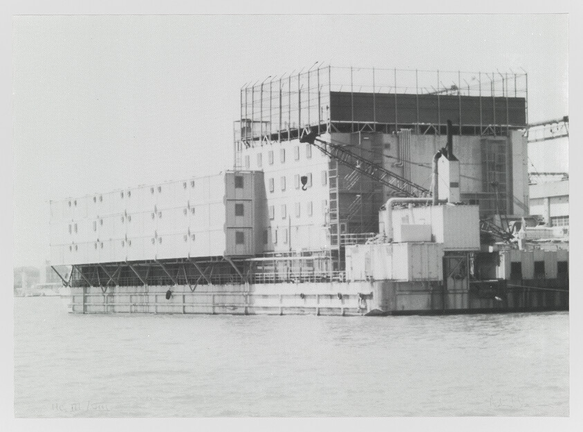 A large industrial dockside building with scaffolding and platforms over calm water.