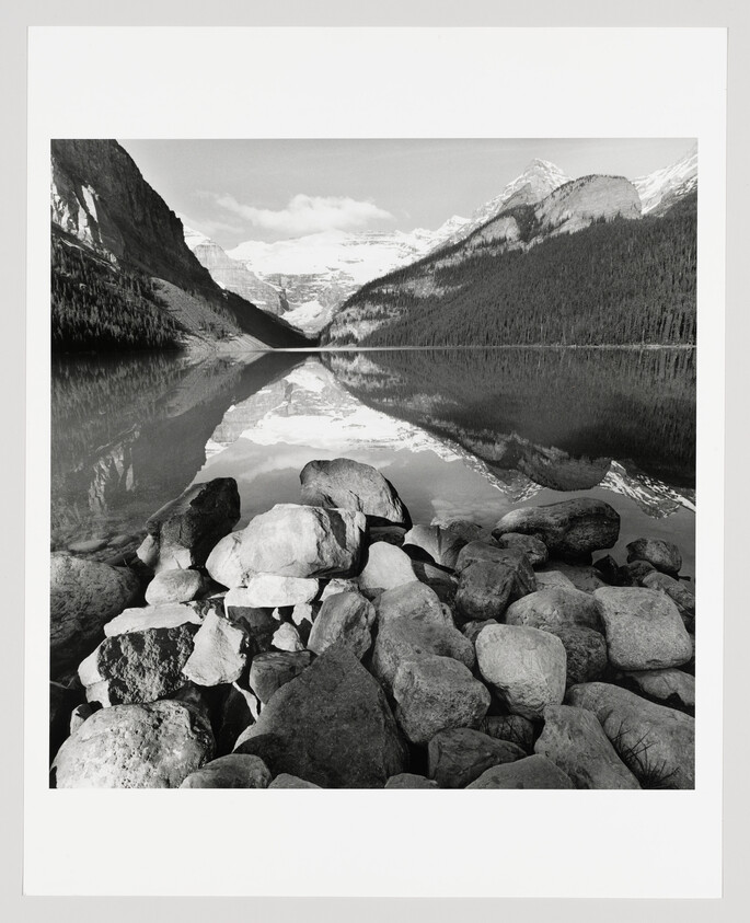 Rocky shoreline leading to a calm mountain lake reflecting snow-covered peaks and forested slopes.