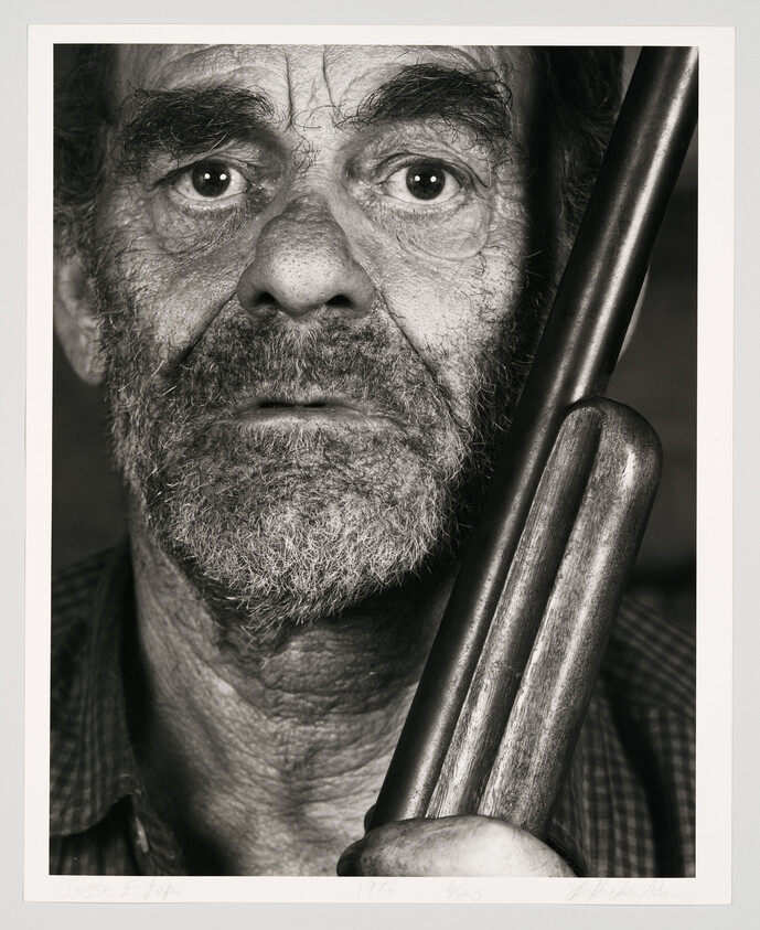 An older bearded man stares at the camera while holding a shotgun barrel close.