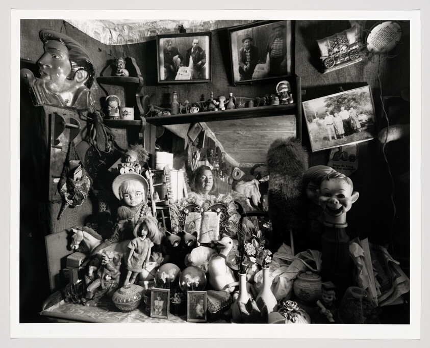 Woman reflected in a mirror surrounded by an assortment of dolls, toys, and small collectibles.