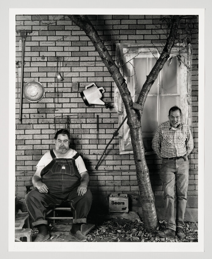 Two men pose on a porch beside a tree, one seated in overalls and one standing smiling.