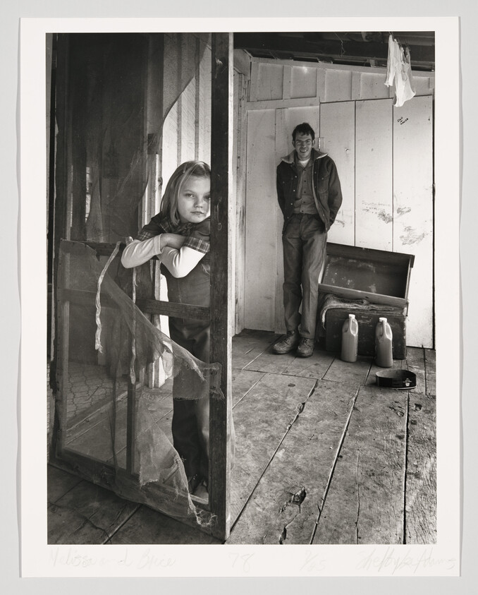 A black and white photograph depicting a young girl leaning on a wooden door frame on the left, looking directly at the camera with a contemplative expression. On the right, a young man stands inside a rustic room, hands in pockets, smiling slightly towards the camera. The room appears worn, with wooden planks for walls and floor, and a couple of jugs and a radio on the floor beside the man. Handwritten text at the bottom of the image includes names and a date, suggesting personal or artistic significance.