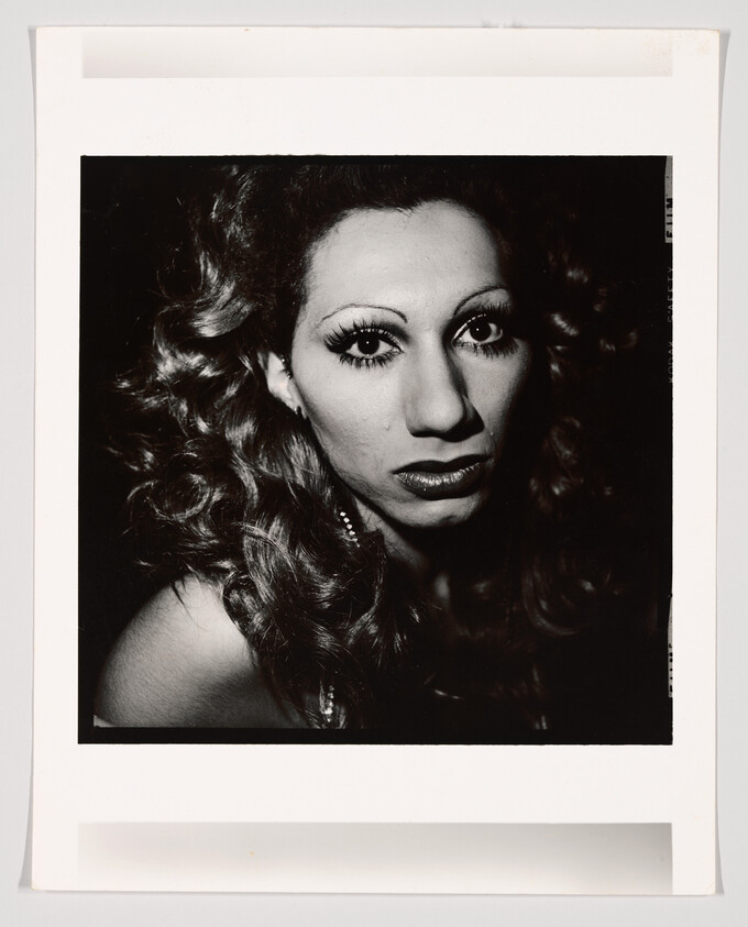 Dramatic black-and-white portrait of a person with heavy eye makeup and curled hair, looking at the camera.