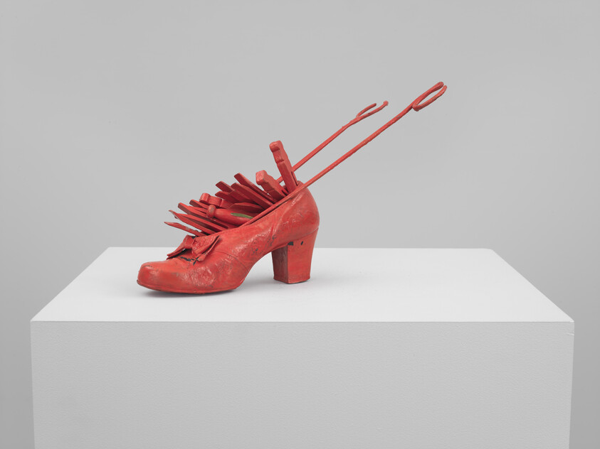 Red sculptural high-heeled shoe with forks and long tongs protruding on a white pedestal.