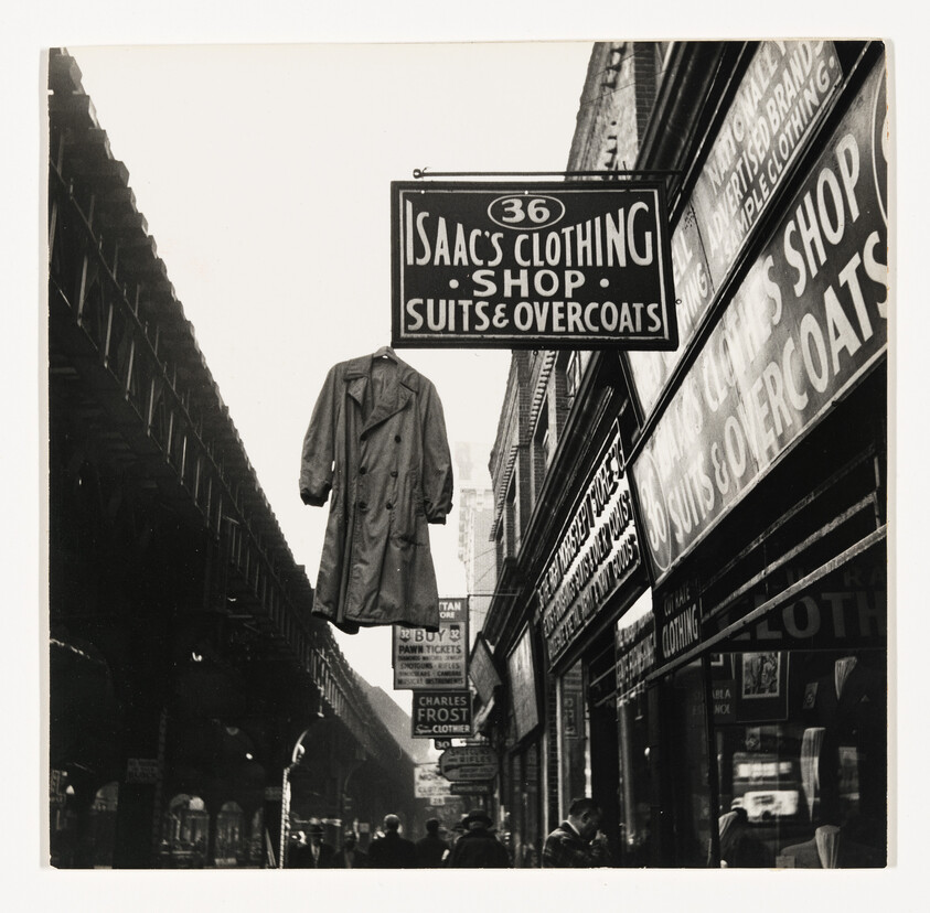 Vintage photo of a street with hanging signs for Isaac's Clothing Shop and other businesses, featuring an overcoat.