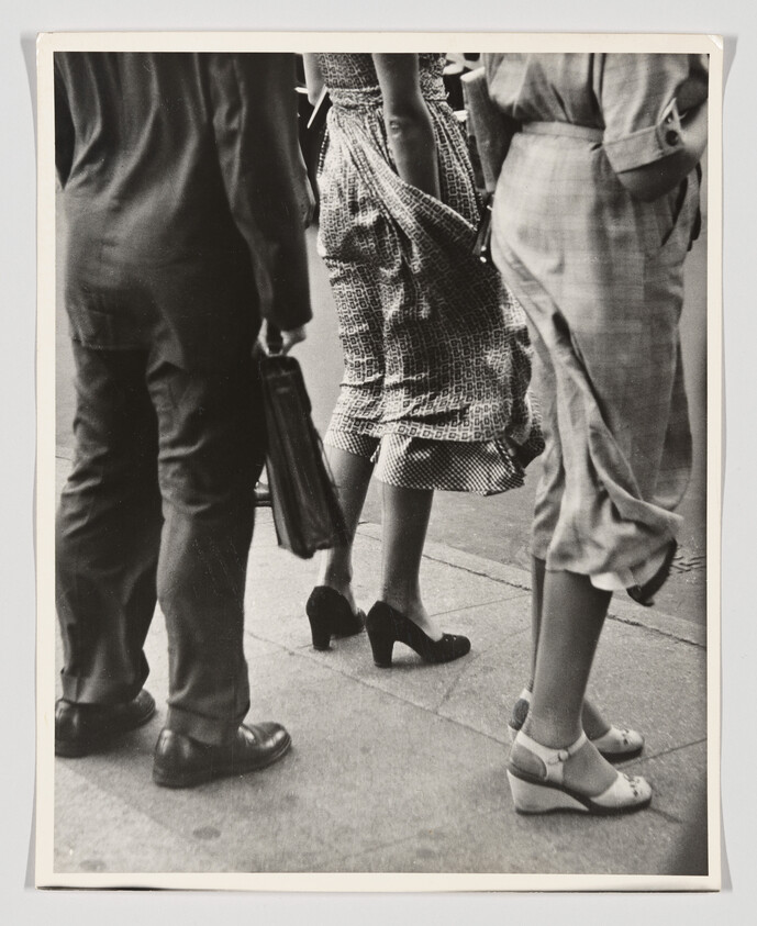 A black and white photograph capturing the lower half of three individuals walking, focusing on their legs and feet. The person on the left is wearing dark trousers and polished shoes, carrying a briefcase. The middle person is dressed in a patterned skirt and heels, while the person on the right is wearing light-colored trousers and two-tone strapped shoes. The setting appears to be an urban sidewalk.