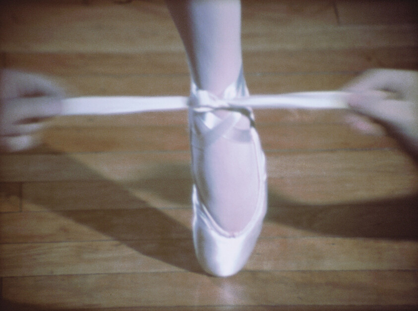 Ballerina ties satin pointe shoe ribbon around foot while standing on a wooden floor.