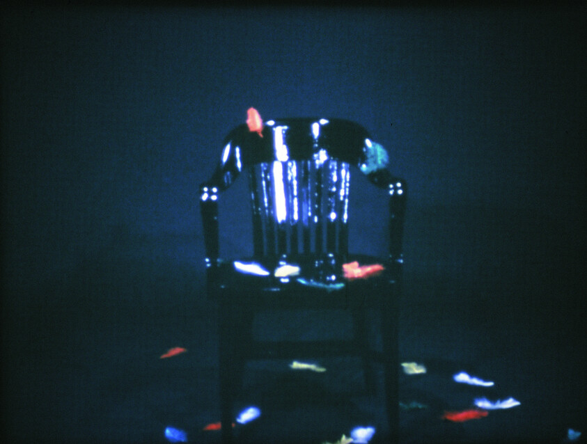 A shiny black wooden chair with colorful feathers scattered on and around it, set against a dark background.