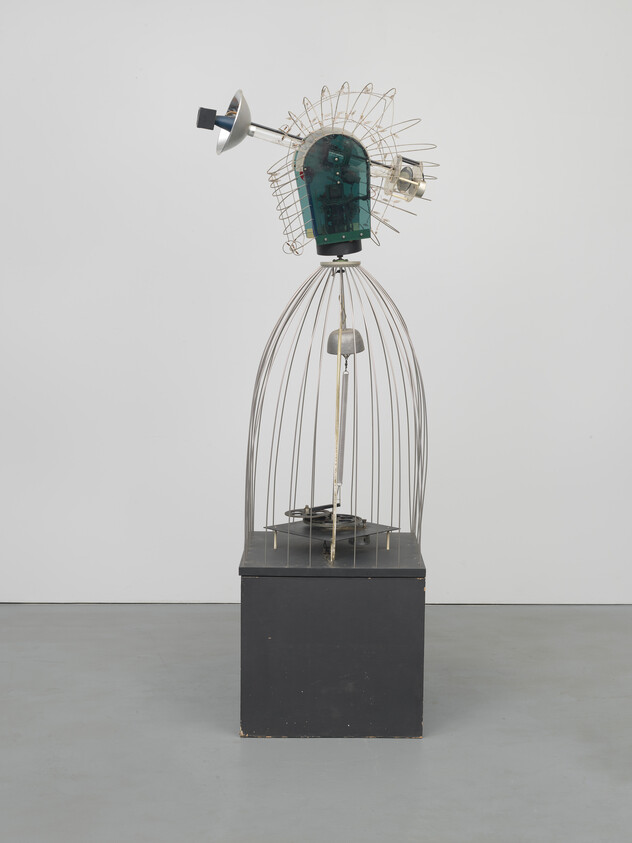 Sculpture of a humanoid figure with a wireframe head and body, electronic components for a face, standing on a black pedestal.