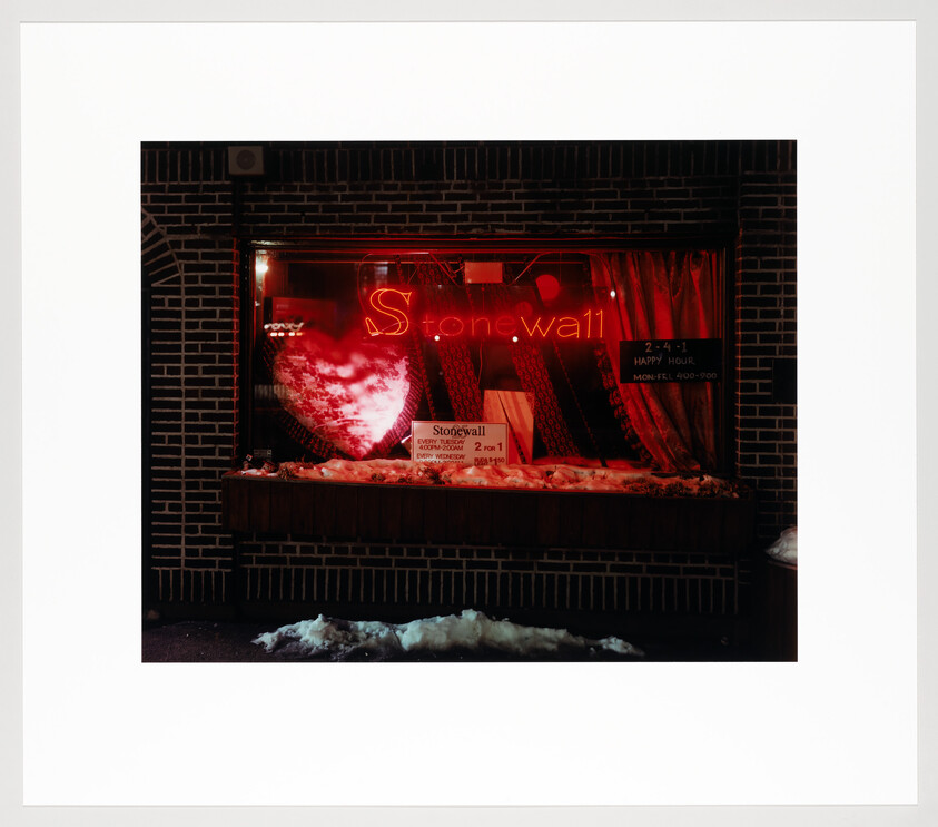 Storefront window lit by red neon sign reading Stonewall above a display, with snow on the sidewalk.