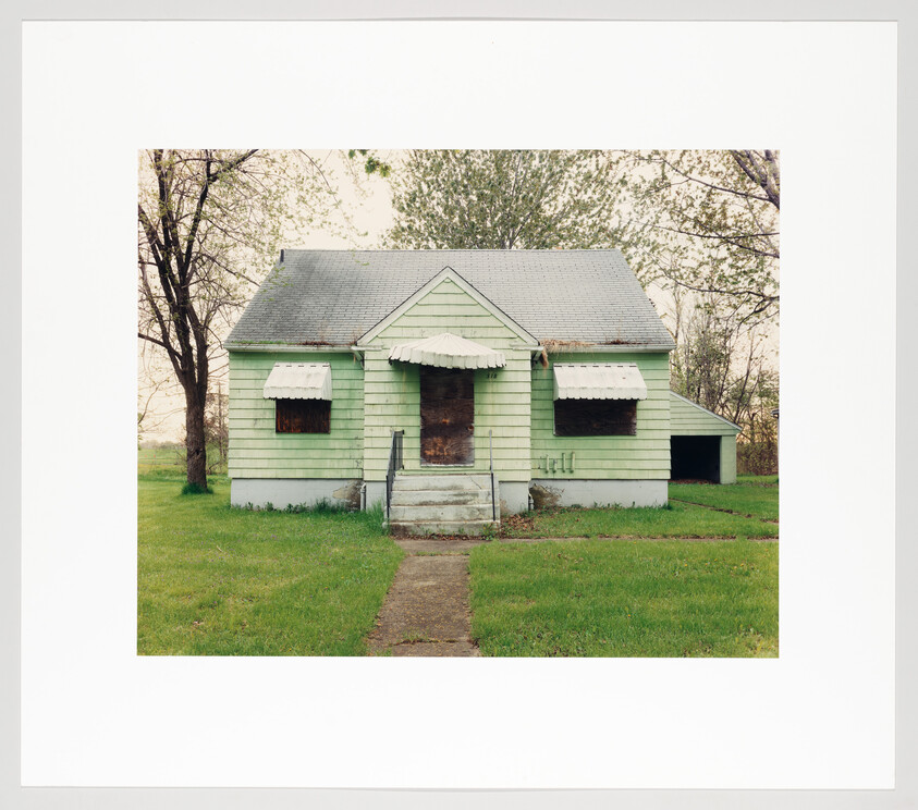 Small pale green house with boarded windows and door and a cracked front path.