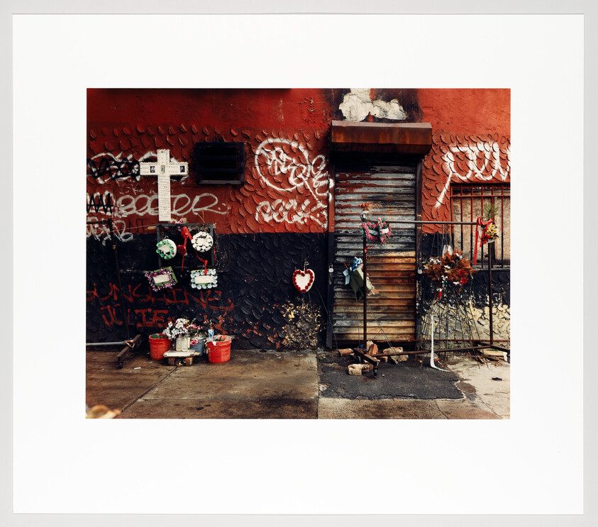 Makeshift street memorial with cross, wreaths, and flowers against a graffiti-covered wall and closed metal door.