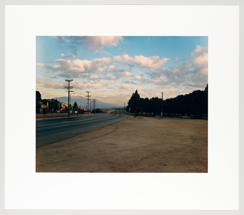 Quiet empty highway with dirt shoulder and telephone poles stretching toward distant mountains under cloudy sky.
