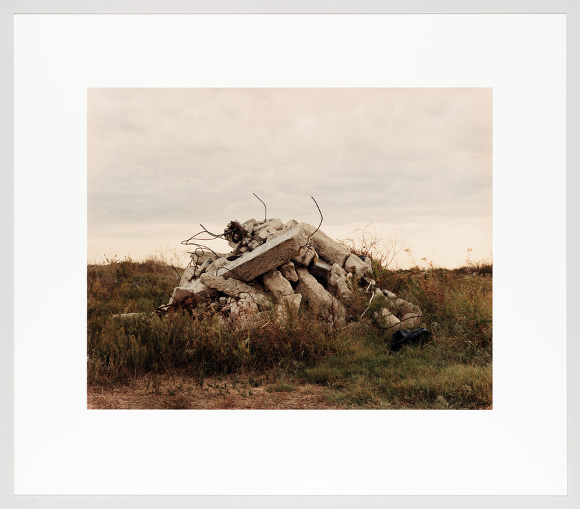 A pile of broken concrete with exposed rebar sits in a grassy field under a cloudy sky.