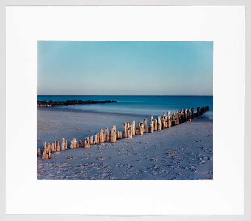 Weathered wooden posts line the sandy beach and stretch diagonally into the calm blue sea.