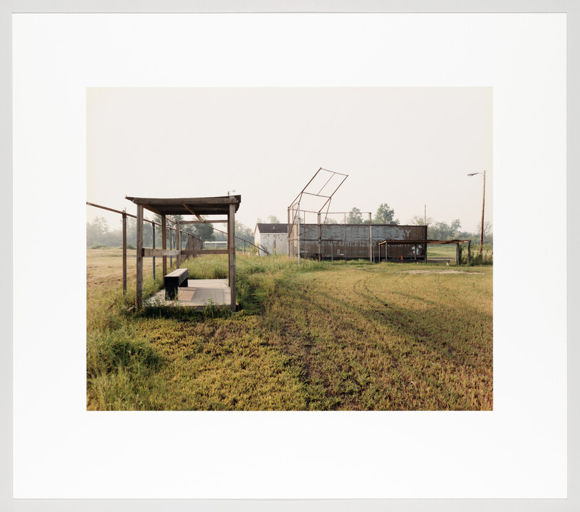 An overgrown, unused baseball field with a weathered dugout bench and broken backstop in the distance.