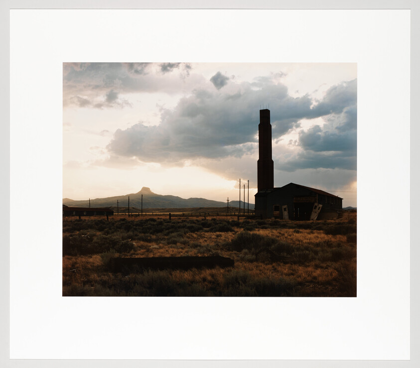 An abandoned industrial building with a tall smokestack stands in grassy plains under a cloudy sunset.
