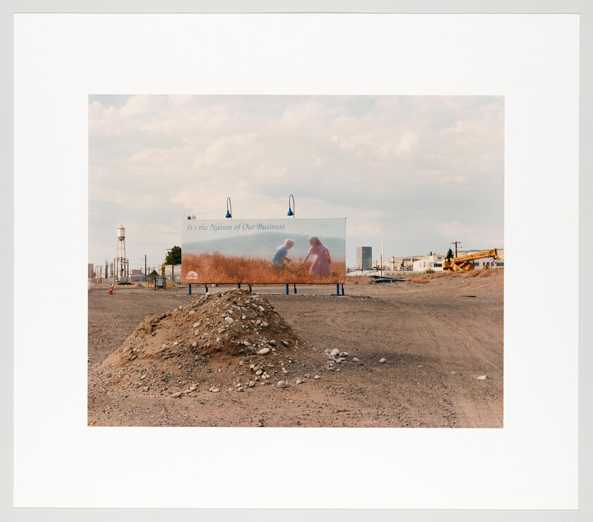 A billboard reading "It's the nature of our business" stands behind a dirt mound in an empty lot.
