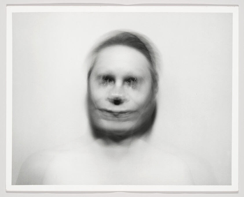 A blurred, pale face with smeared dark eyes and mouth against a plain background.