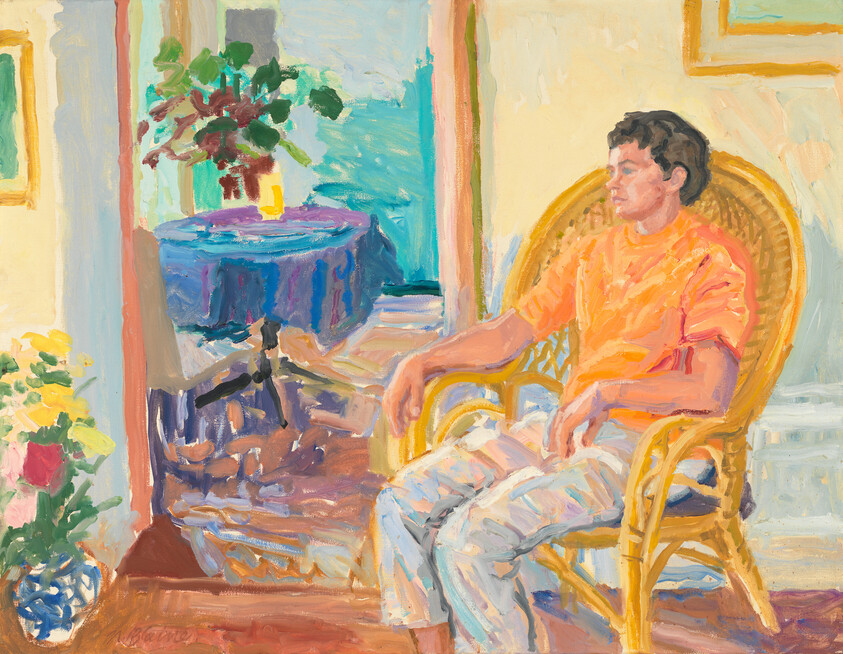 A young man in an orange shirt sits relaxed in a wicker chair looking toward a bright room.