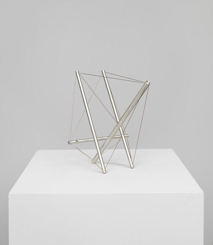 A small geometric metal sculpture of intersecting rods held together with thin wires on a pedestal.
